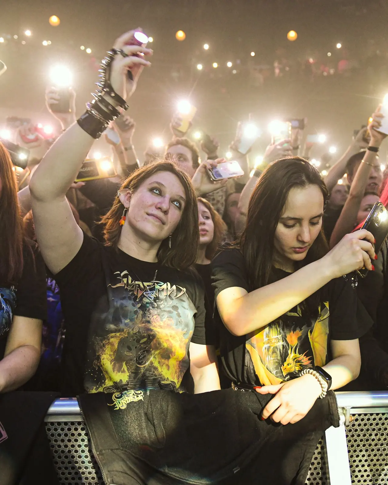 Two fans at a concert barrier holding up their phones in the stage lights
